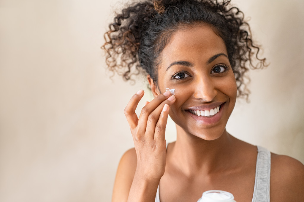 beautiful curly haired woman smiling while applying cream in cheek